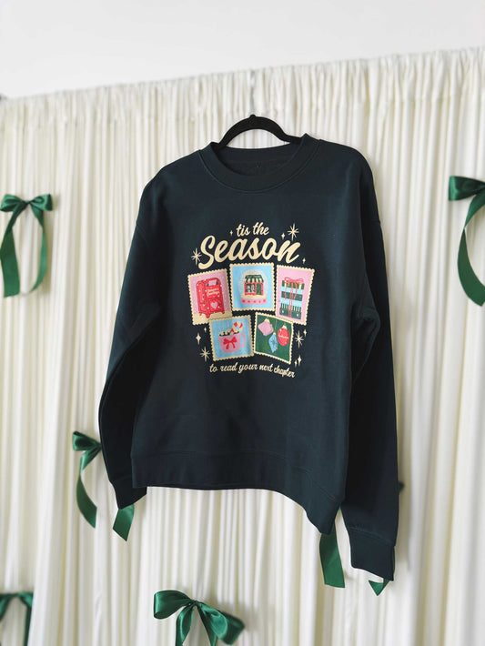Next Chapter Holiday Crewneck. A Dark green sweatshirt with a festive bookish Christmas design and 'tis the season' text in a casual indoor setting.