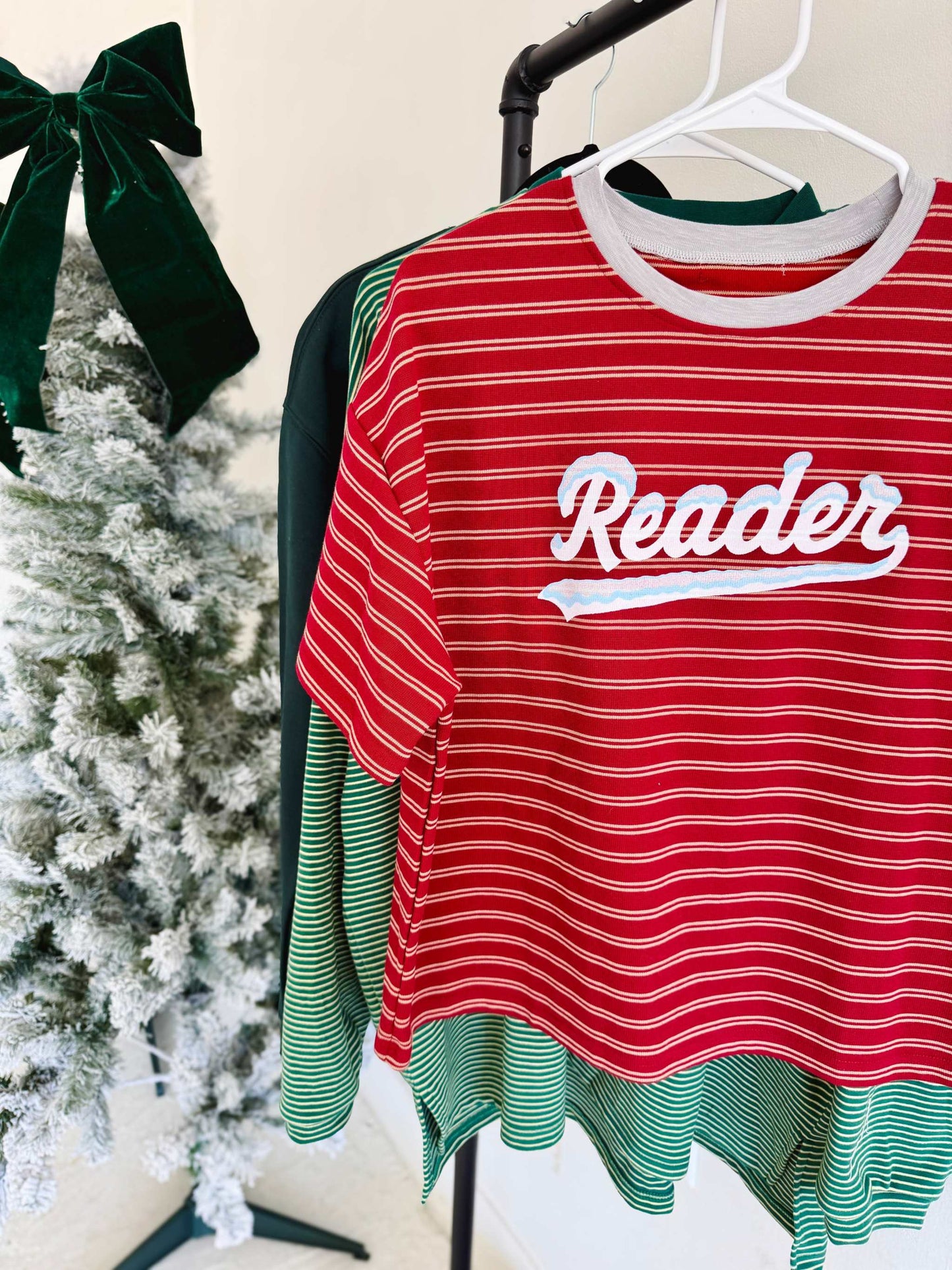 Red Snowy Reader Striped Boxy Top t-shirt with snowcapped 'Reader' text on a hanger near a Christmas tree.