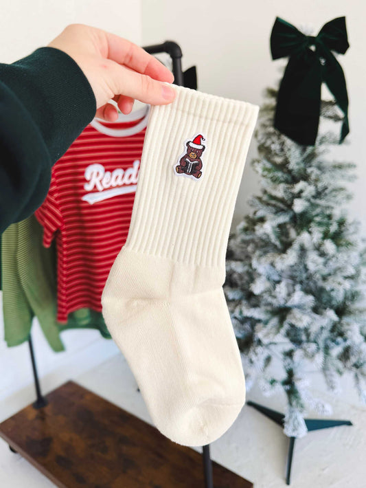 Santa Book Bear Socks are held up in front of the camera with Christmas themed props in the background. The socks have a holiday bear with a santa hat holding a book.
