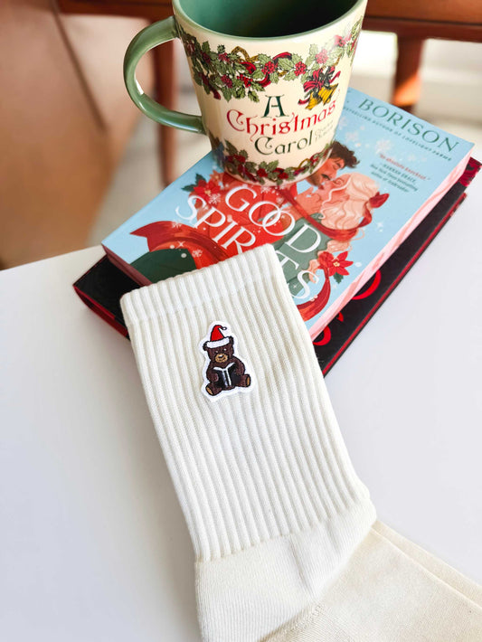 Santa Book Bear Socks are propped up near books and a mug on a coffee table. The socks feature a holiday themed bear holding a book.