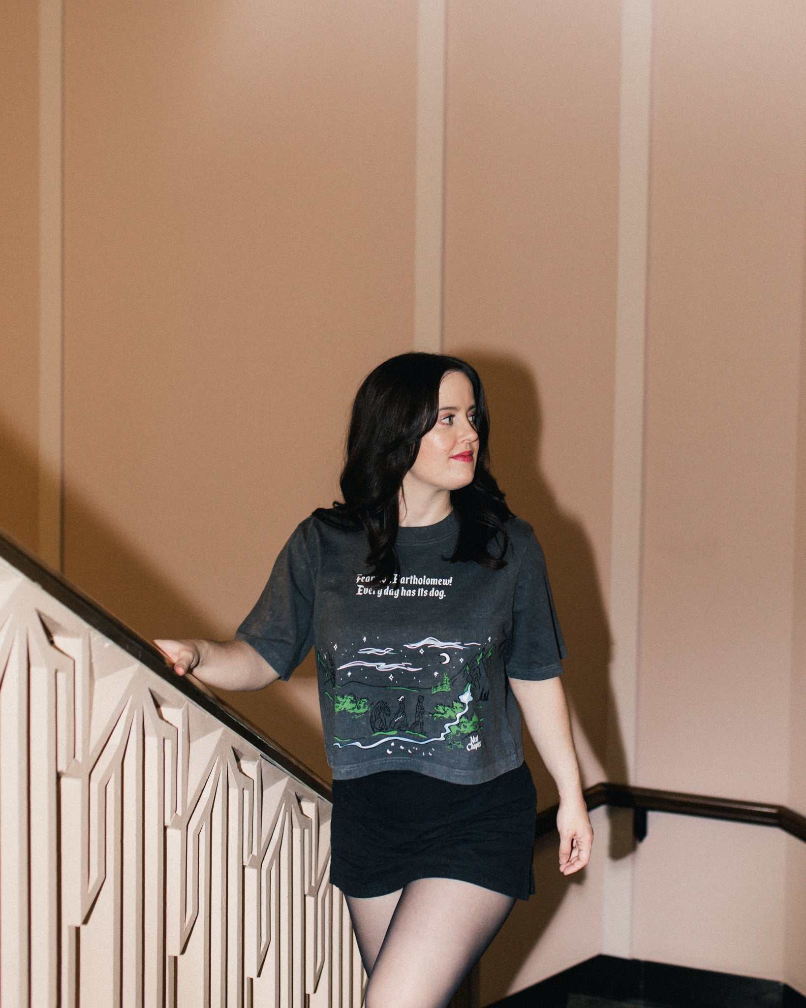 Person wearing The Unlikely Trio Tee that is dark gray and cropped with a graphic design and text that reads 'Fear not, Bartholomew! Every day has its dog!' Inspired by The Knight and the Moth by Rachel Gillig.