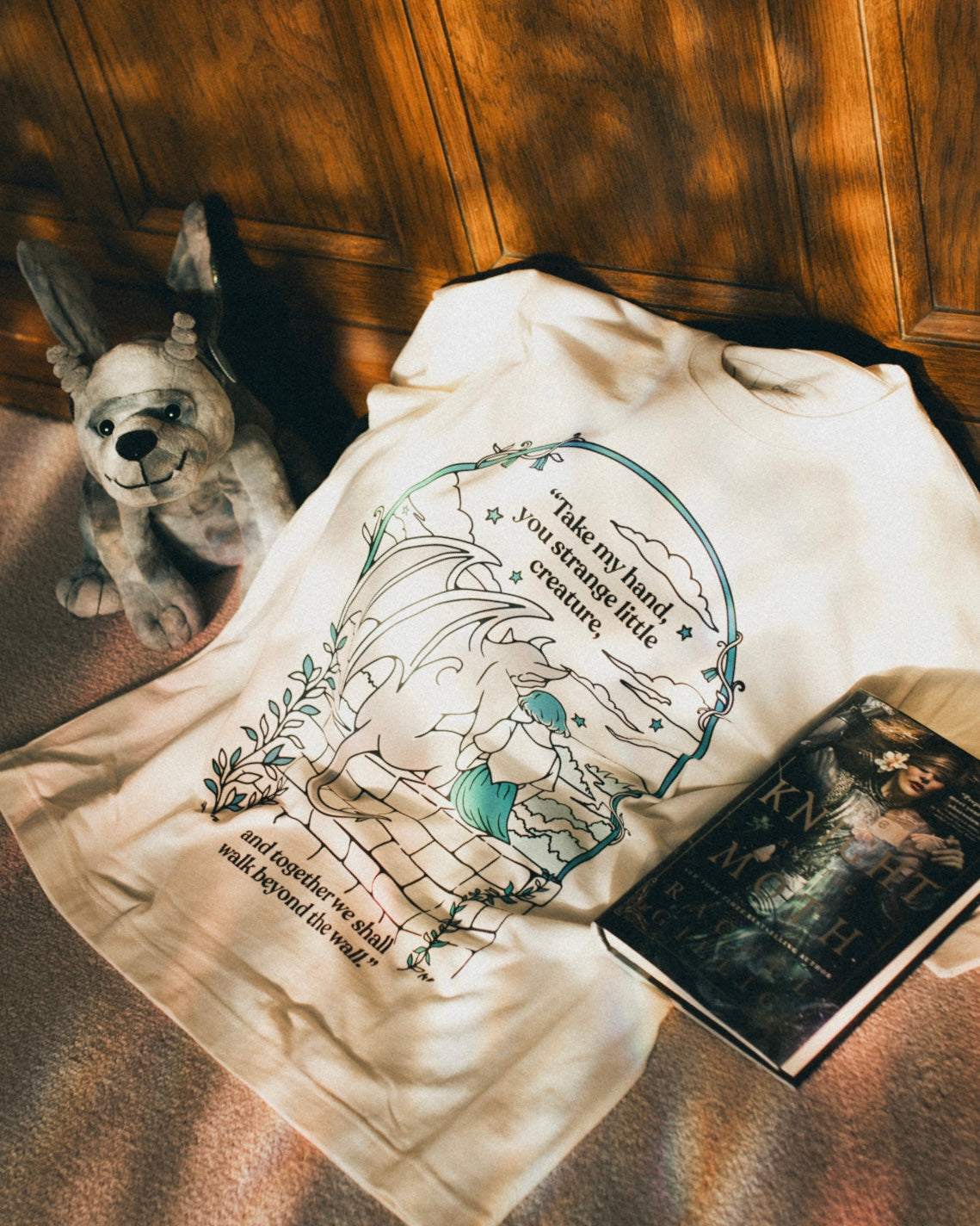 Ivory Strange Little Creatures Tee with graphic design inspired by The Knight and the Moth by Rachel Gillig sitting next to a Gargoyle Plushie and book.
