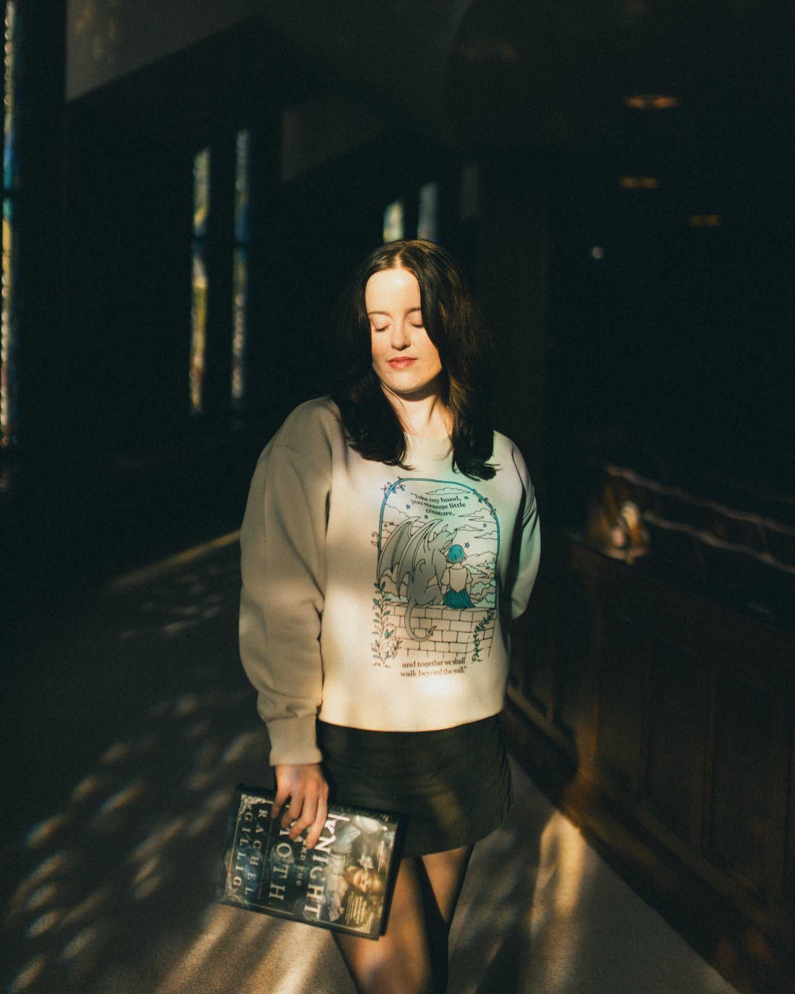 Woman wearing an ivory strange little creatures crewneck sweatshirt with a graphic design inspired by The Knight and the Moth by Rachel Gillig.