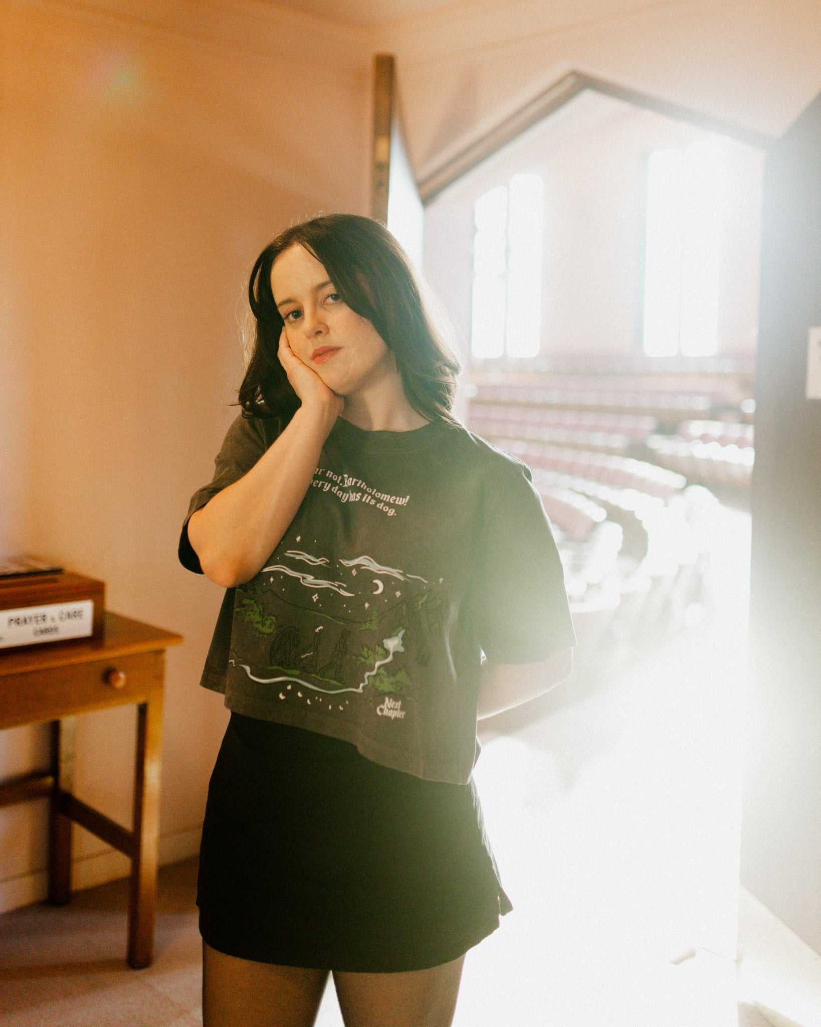 Person wearing The Unlikely Trio Tee that is dark gray and cropped with a graphic design and text that reads 'Fear not, Bartholomew! Every day has its dog!' Inspired by The Knight and the Moth by Rachel Gillig.