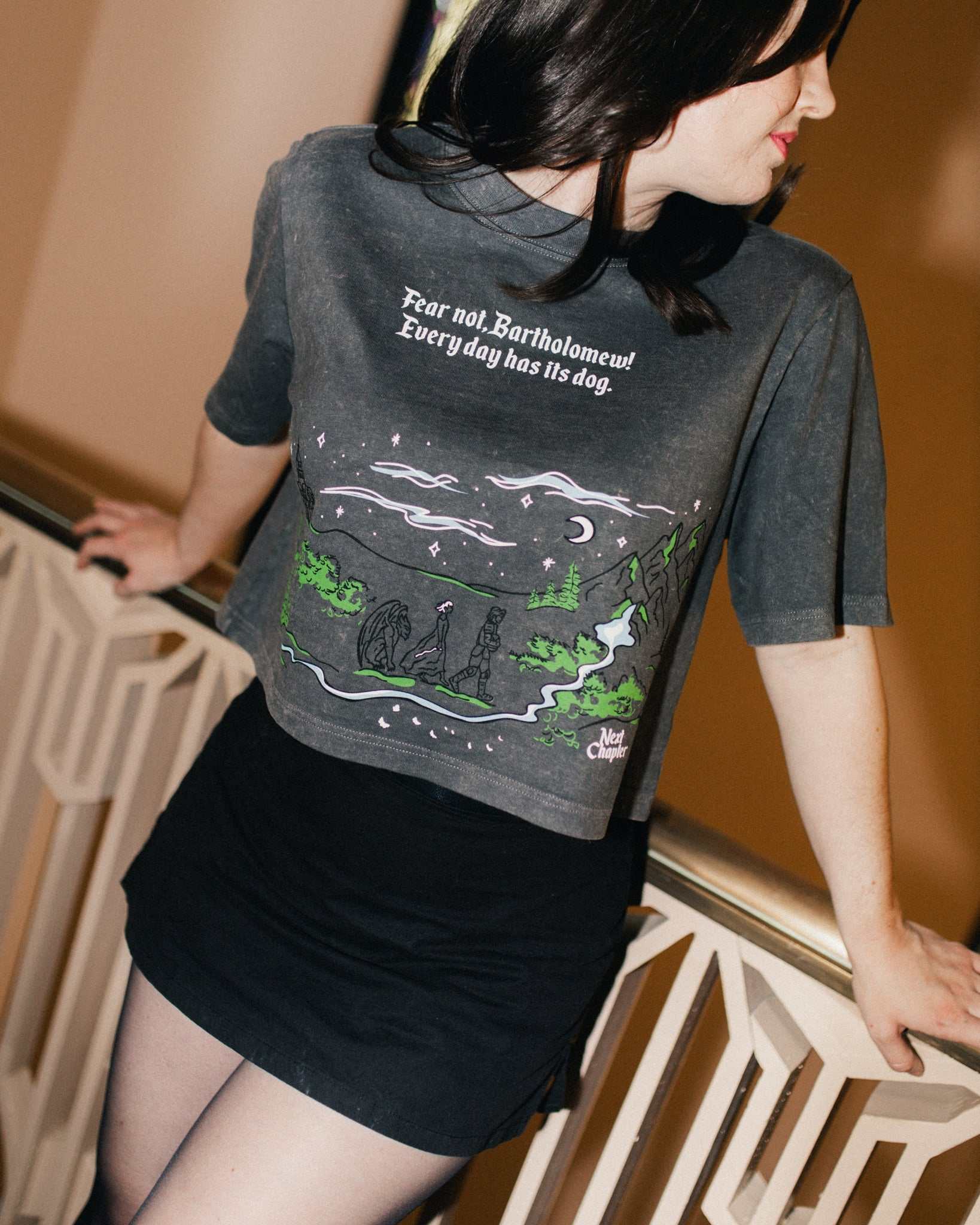 Person wearing The Unlikely Trio Tee that is dark gray and cropped with a graphic design and text that reads 'Fear not, Bartholomew! Every day has its dog!' Inspired by The Knight and the Moth by Rachel Gillig.