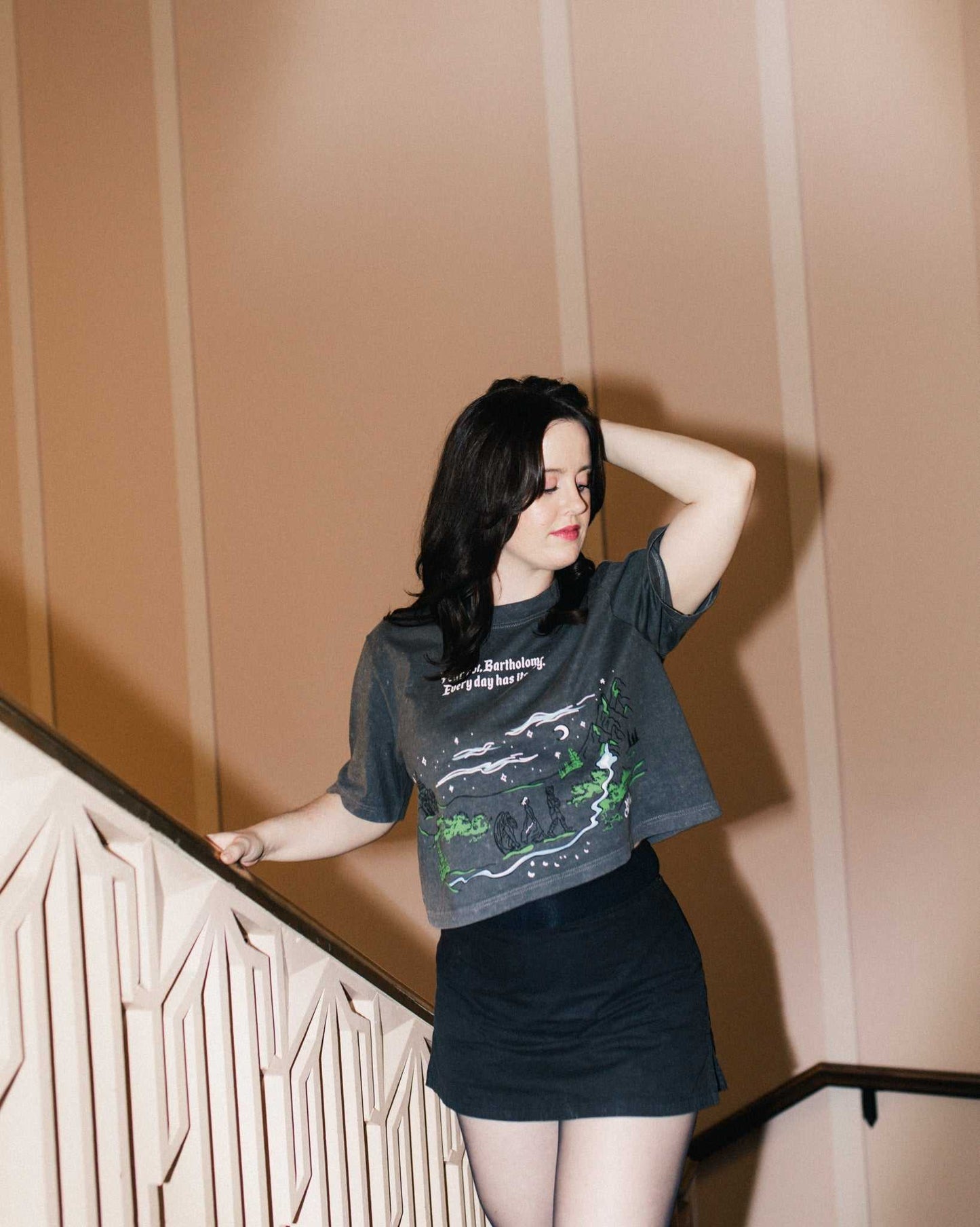 Person wearing The Unlikely Trio Tee that is dark gray and cropped with a graphic design and text that reads 'Fear not, Bartholomew! Every day has its dog!' Inspired by The Knight and the Moth by Rachel Gillig.