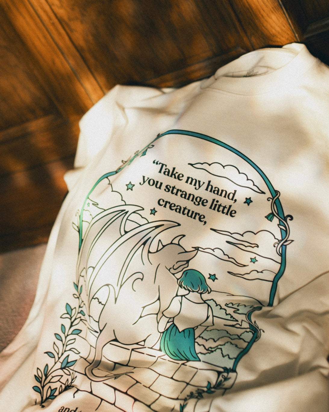 Ivory Strange Little Creatures Tee with a graphic design featuring Gargoyle and Sybil Delling from The Knight and the Moth by Rachel Gillig and text saying 'Take my hand, you strange little creature'.