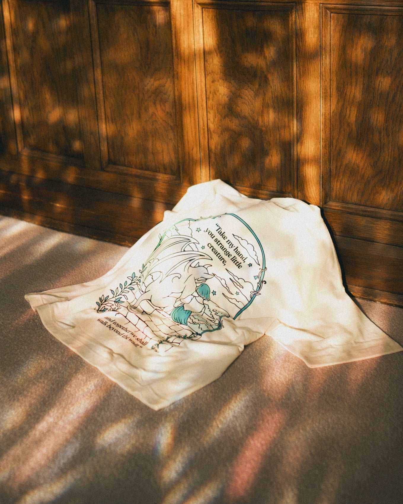 Ivory Strange Little Creatures Tee with a graphic design featuring Gargoyle and Sybil Delling from The Knight and the Moth by Rachel Gillig and text saying 'Take my hand, you strange little creature, and together we shall walk beyond the wall.'