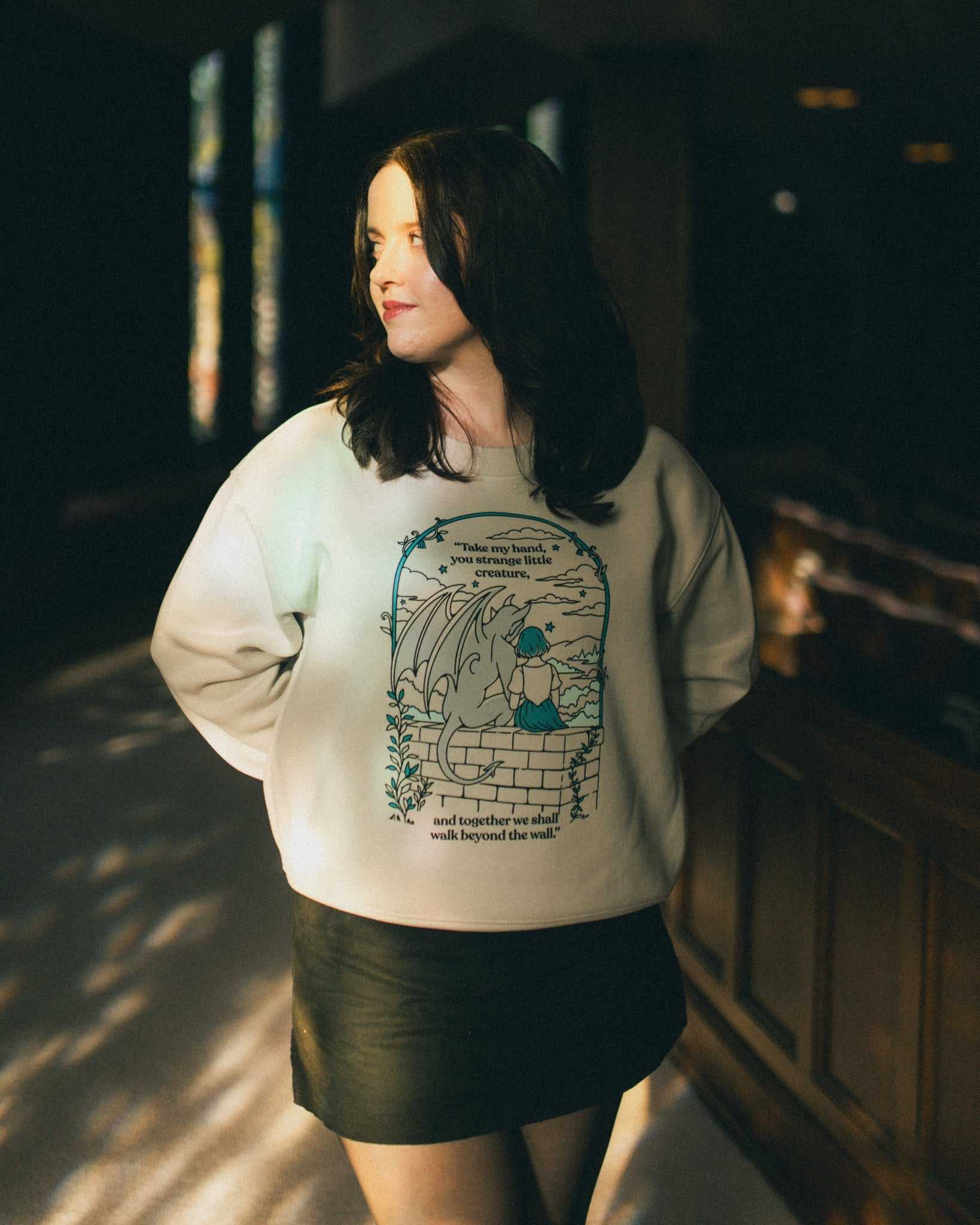Woman wearing an ivory strange little creatures crewneck sweatshirt with a graphic design inspired by The Knight and the Moth by Rachel Gillig.