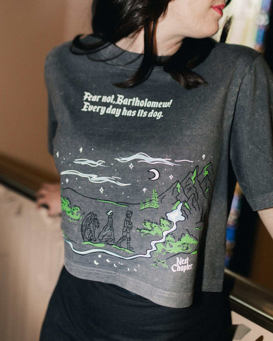 Person wearing The Unlikely Trio Tee that is dark gray and cropped with a graphic design and text that reads 'Fear not, Bartholomew! Every day has its dog!' Inspired by The Knight and the Moth by Rachel Gillig.