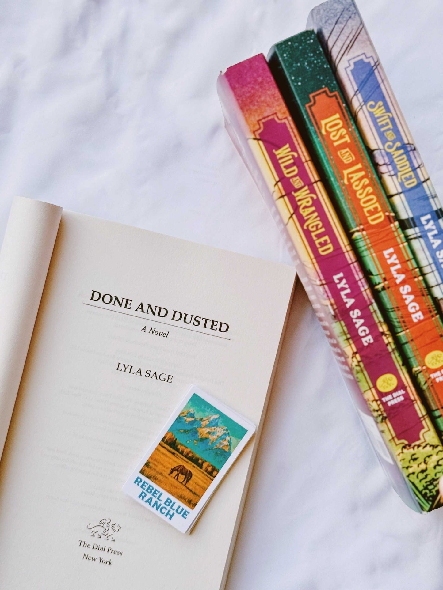 A Rebel Blue Ranch Polaroid Sticker from 'Done and Dusted' by Lyla Sage with a horse and landscape design, placed on top of other books.