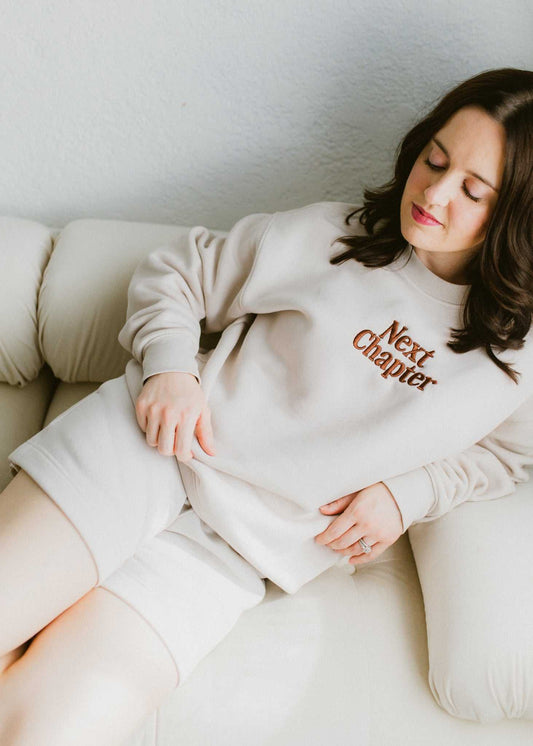 Woman wearing ivory Next Chapter Crewneck sweatshirt with chocolate brown print, relaxing on a couch