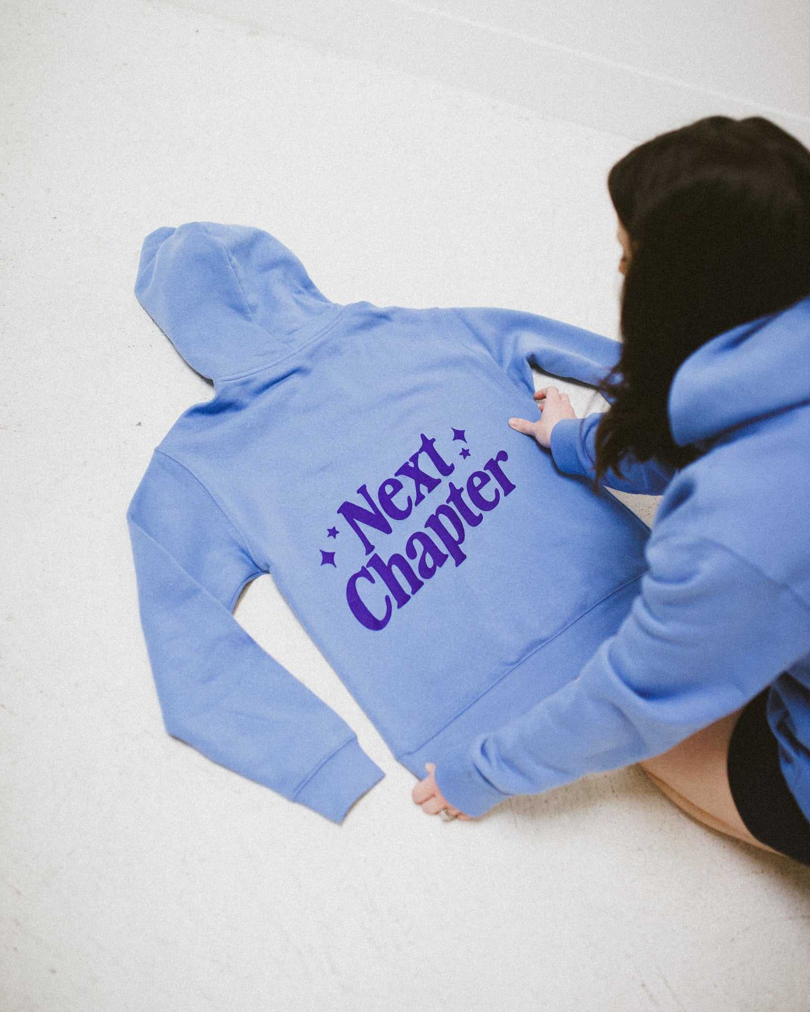 Person wearing a blue Next Chapter hoodie with 'Next Chapter' text on a light background. Next Chapter is a bookish small business that offers officially licensed book merch, including Rachel Gillig merch and Lyla Sage merch.