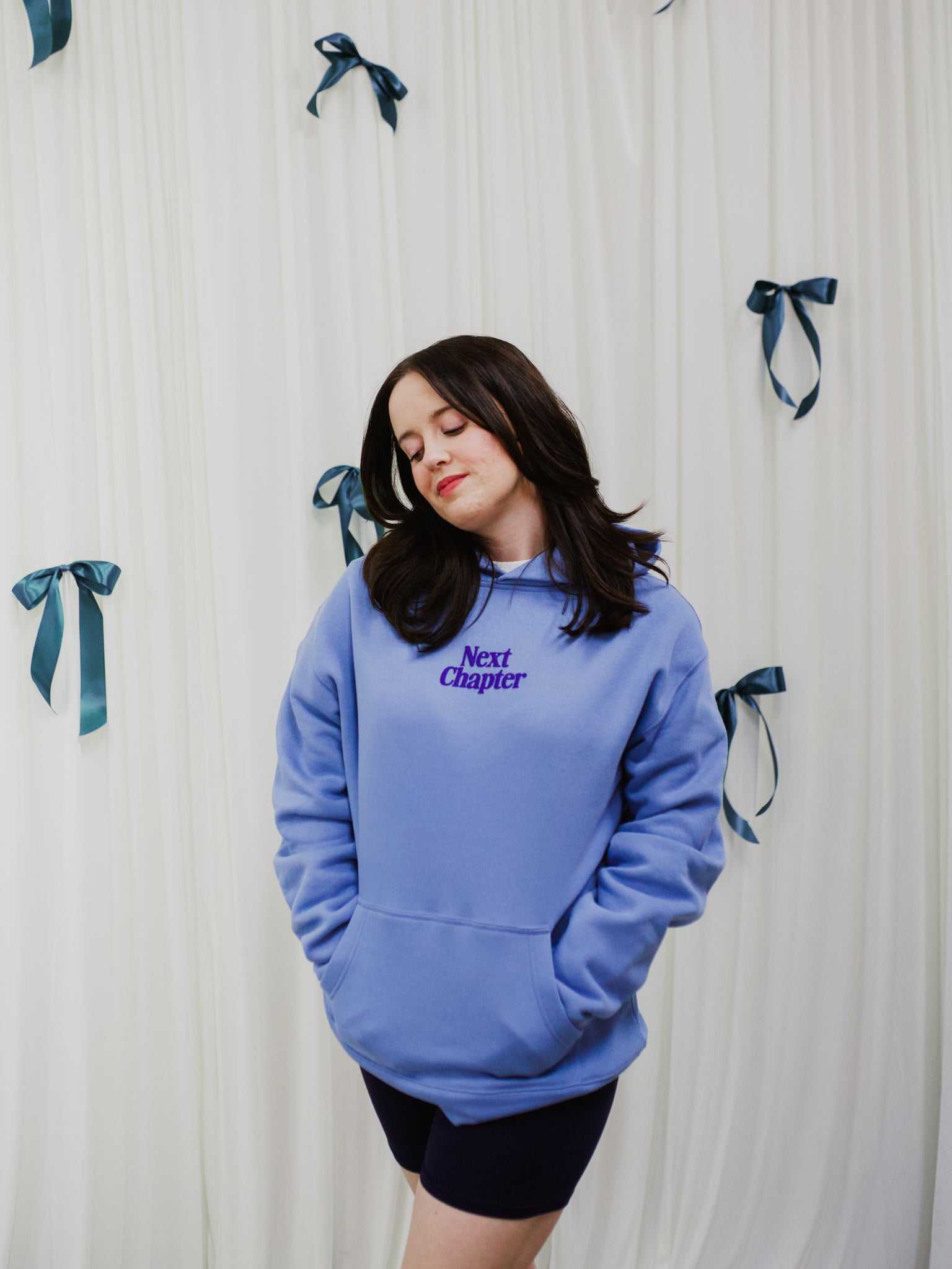 Person wearing a blue Next Chapter hoodie with 'Next Chapter' text against a white curtain background. Next Chapter is a bookish small business that offers officially licensed book merch, including Rachel Gillig merch and Lyla Sage merch.