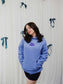 Person wearing a blue Next Chapter hoodie with 'Next Chapter' text against a white curtain background. Next Chapter is a bookish small business that offers officially licensed book merch, including Rachel Gillig merch and Lyla Sage merch.