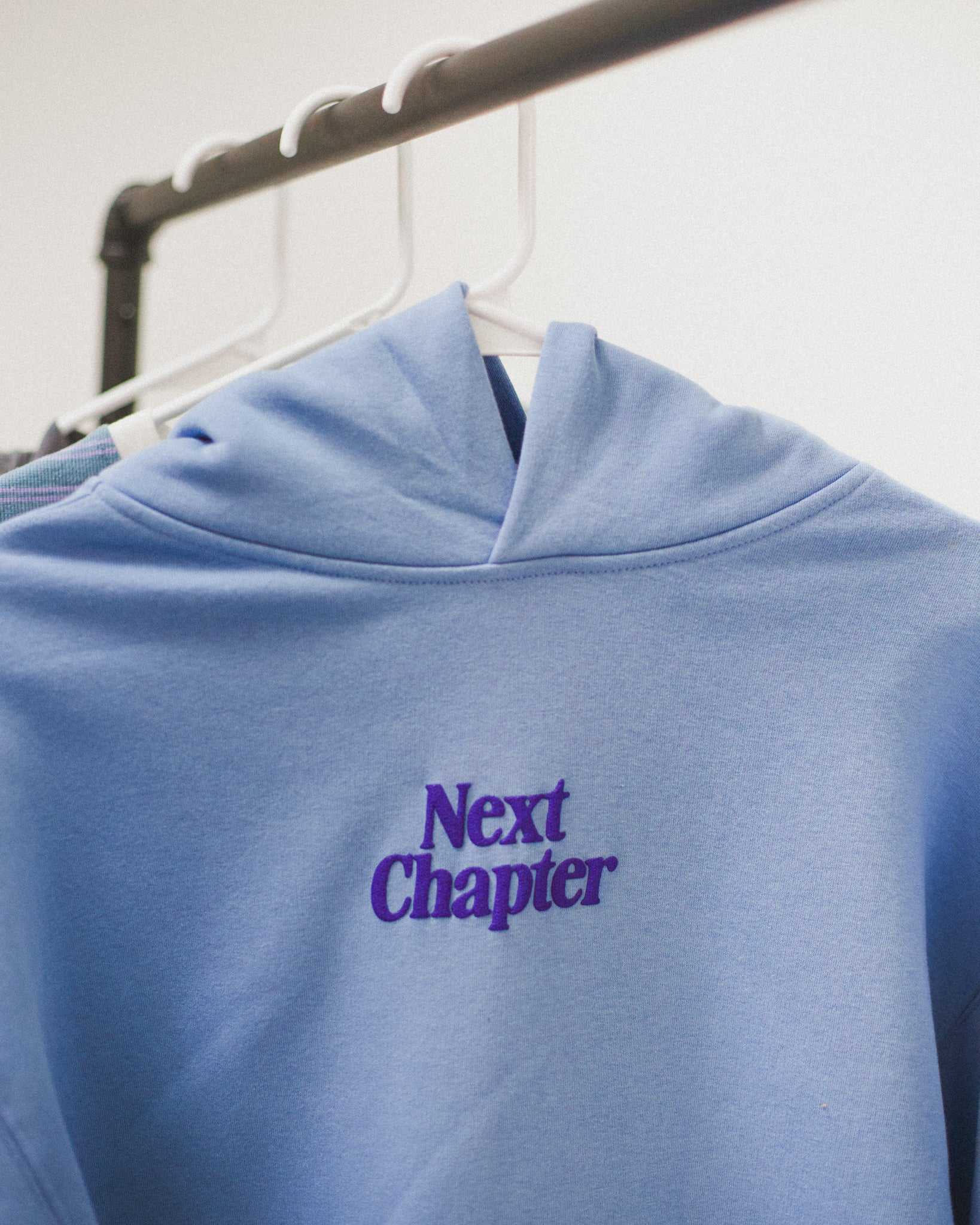 Blue Next Chapter hoodie with 'Next Chapter' puff print on a hanger against a white background. Next Chapter is a bookish small business that offers officially licensed book merch, including Rachel Gillig merch and Lyla Sage merch.