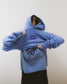 Person wearing a blue Next Chapter hoodie with 'Next Chapter' puff print text on the back against a plain background. Next Chapter is a bookish small business that offers officially licensed book merch, including Rachel Gillig merch and Lyla Sage merch.