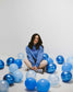Woman sitting on the floor wearing a Next Chapter hoodie, surrounded by blue and white balloons against a plain background. Next Chapter is a bookish small business that offers officially licensed book merch, including Rachel Gillig merch and Lyla Sage merch.