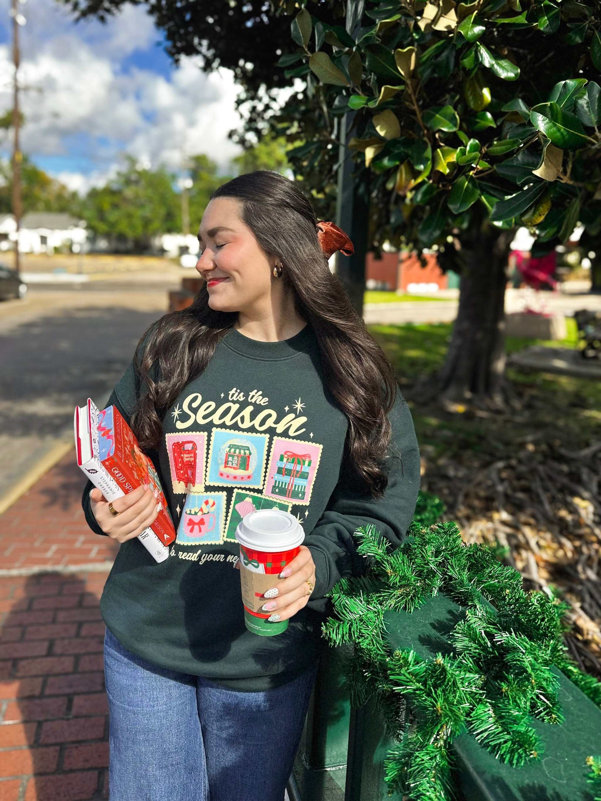 Woman wearing a festive Next Chapter Holiday Crewneck Sweatshirt with bookish Christmas-themed graphics, holding a coffee cup, standing outdoors.