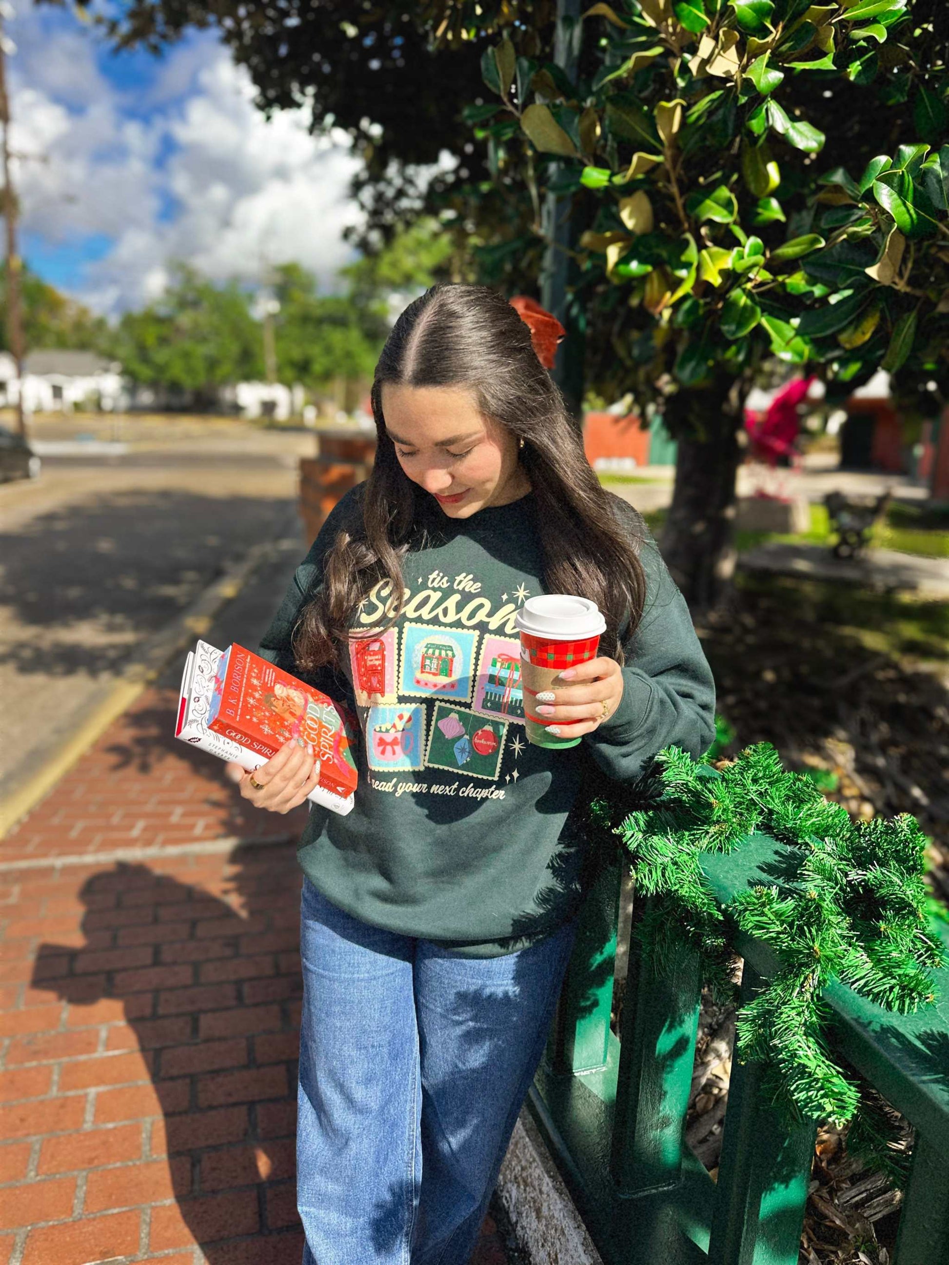 Woman wearing a festive Next Chapter Holiday Crewneck Sweatshirt with bookish Christmas-themed graphics, holding a coffee cup, standing outdoors.