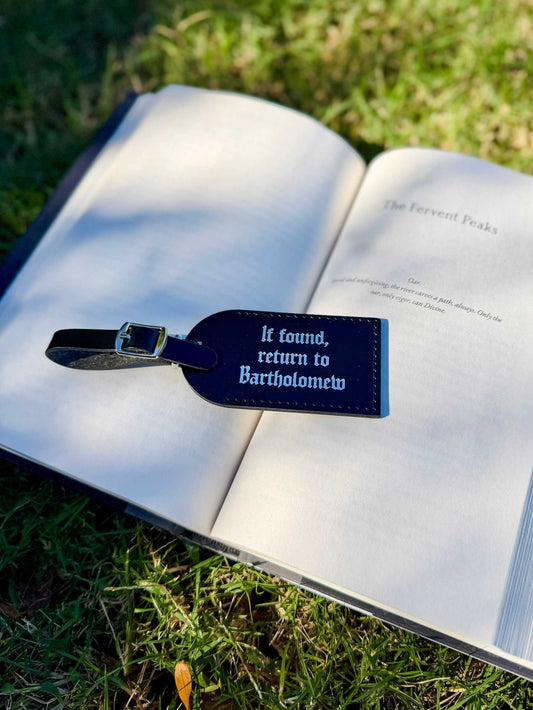 The Bartholomew luggage tags lie on a book outdoors. It reads "If found, return to Bartholomew" and is inspired by The Knight and the Moth by Rachel Gillig.