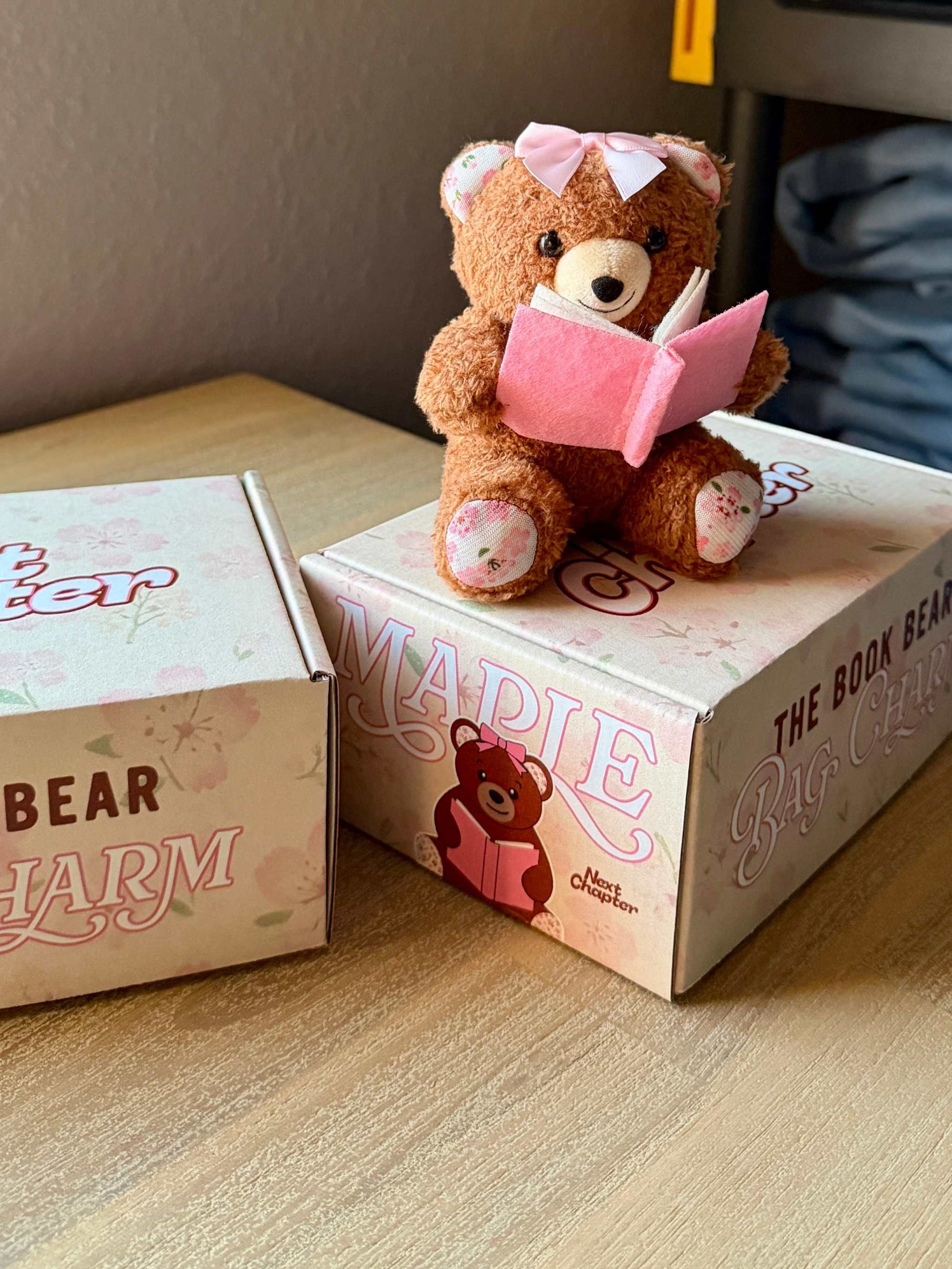 Teddy bear with a pink book on a box labeled 'Maple' on a wooden surface. The Book Bear Plushie Bag Charm from Next Chapter is a part of their "book buddies" collection. Maple, the bookish teddy bear, can be used as a bag charm or bookshelf decor.