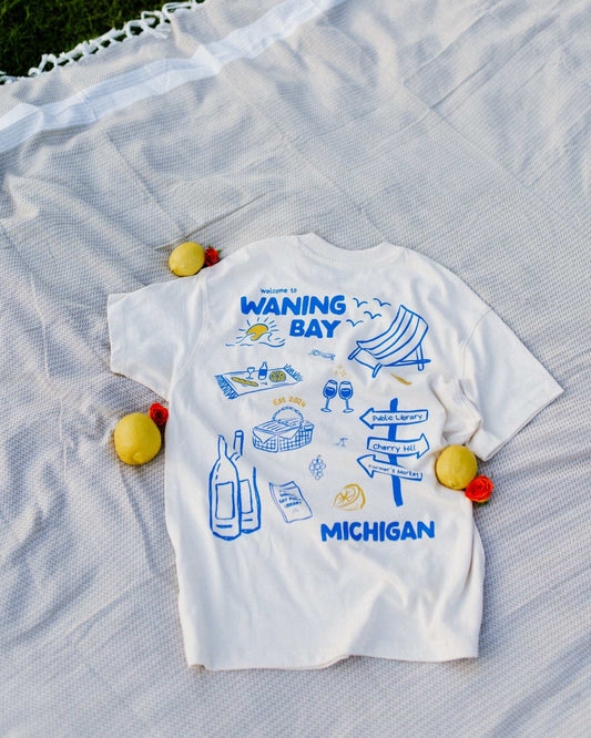 An oversized, boxy t-shirt, the Funny Story Tee is inspired by the book Funny Story by Emily Henry. The item is an ivory/cream color with a royal blue and gold design on the front and back.