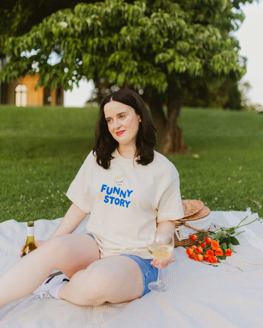 An oversized, boxy t-shirt, the Funny Story Tee is inspired by the book Funny Story by Emily Henry. The item is an ivory/cream color with a royal blue and gold design on the front and back.