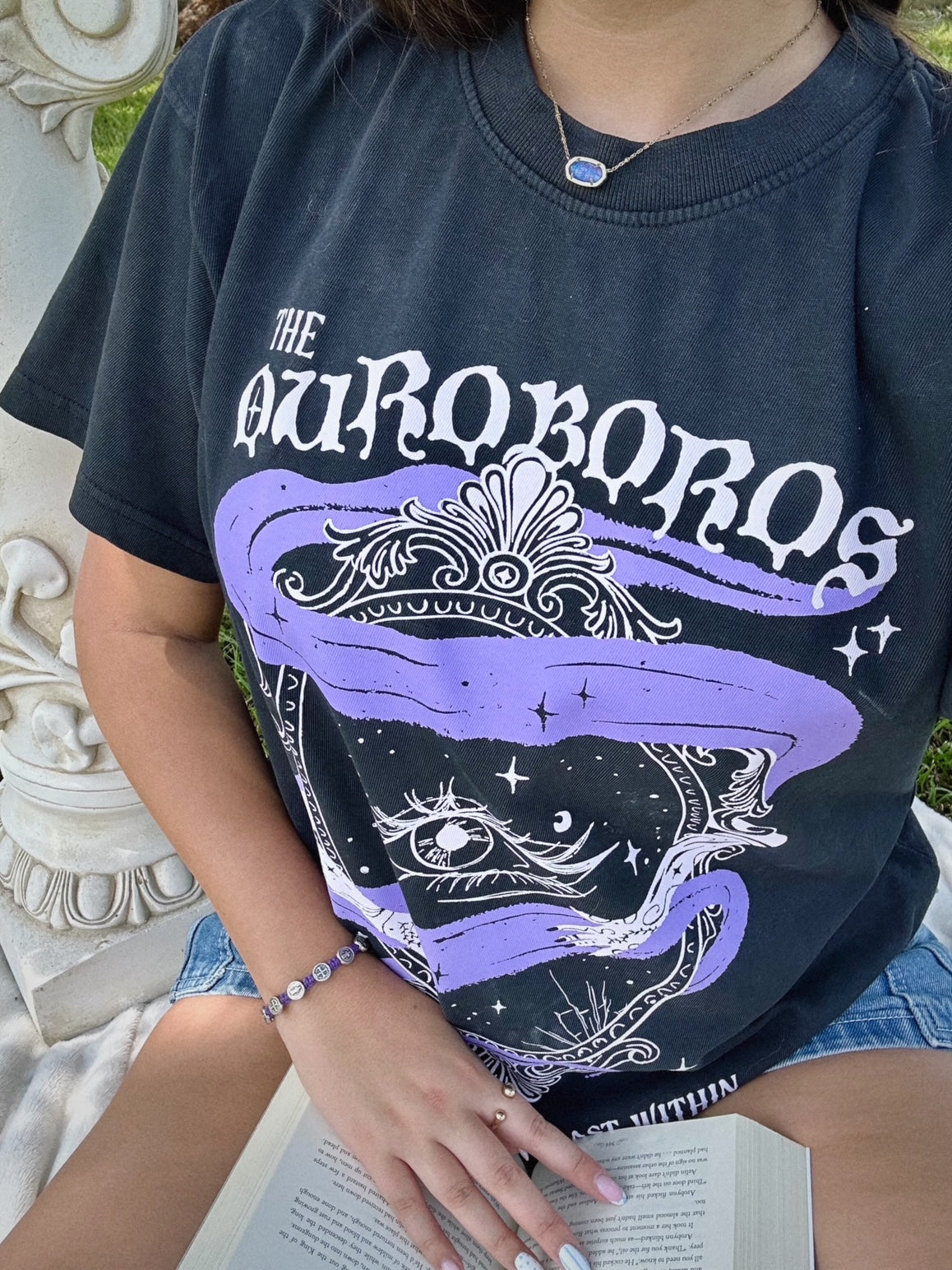 Person wearing a black t-shirt with 'The Ouroboros' design outdoors. A black graphic tee with 'The Ourororos' design featuring an eye, mirror, and purple mist. Inspired by the A Court of Thorns of Roses book series by Sarah J. Maas.