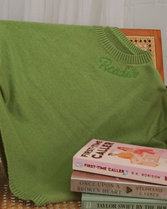 Cozy green Reader Knit Sweater for book lovers draped over a chair with books stacked on it against a white curtain background. The word 'reader' is embroidered along the collar.