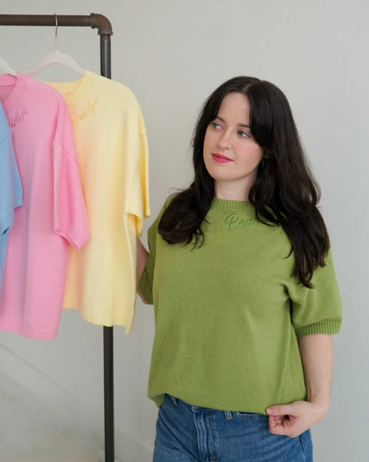 Woman wearing a green Reader Knit Sweater with 'reader' embroidered along the collar, standing next to a rack of colorful sweaters.