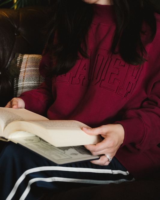The Reader Mock Neck sweatshirt from Next Chapter is a beautiful berry, dark magenta color with an embroidered 'reader' design across the front. It's the perfect cozy bookish sweater for readers and book lovers.