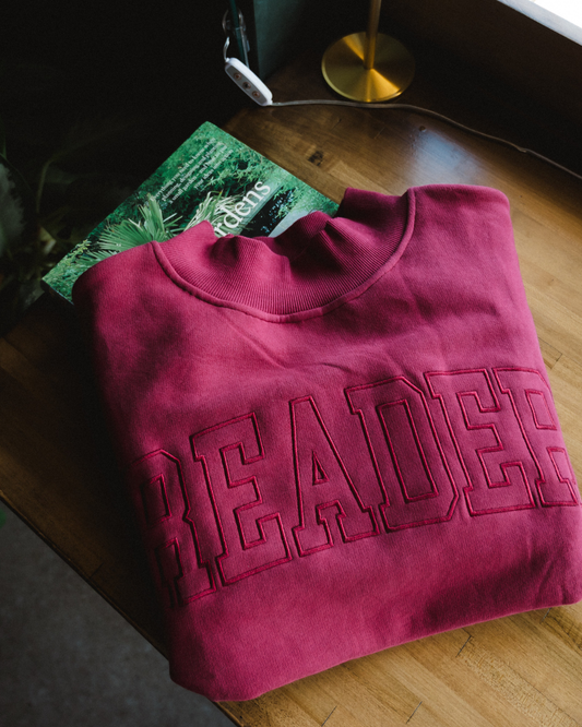 The Reader Mock Neck sweatshirt from Next Chapter is a beautiful berry, dark magenta color with an embroidered 'reader' design across the front. It's the perfect cozy bookish sweater for readers and book lovers.