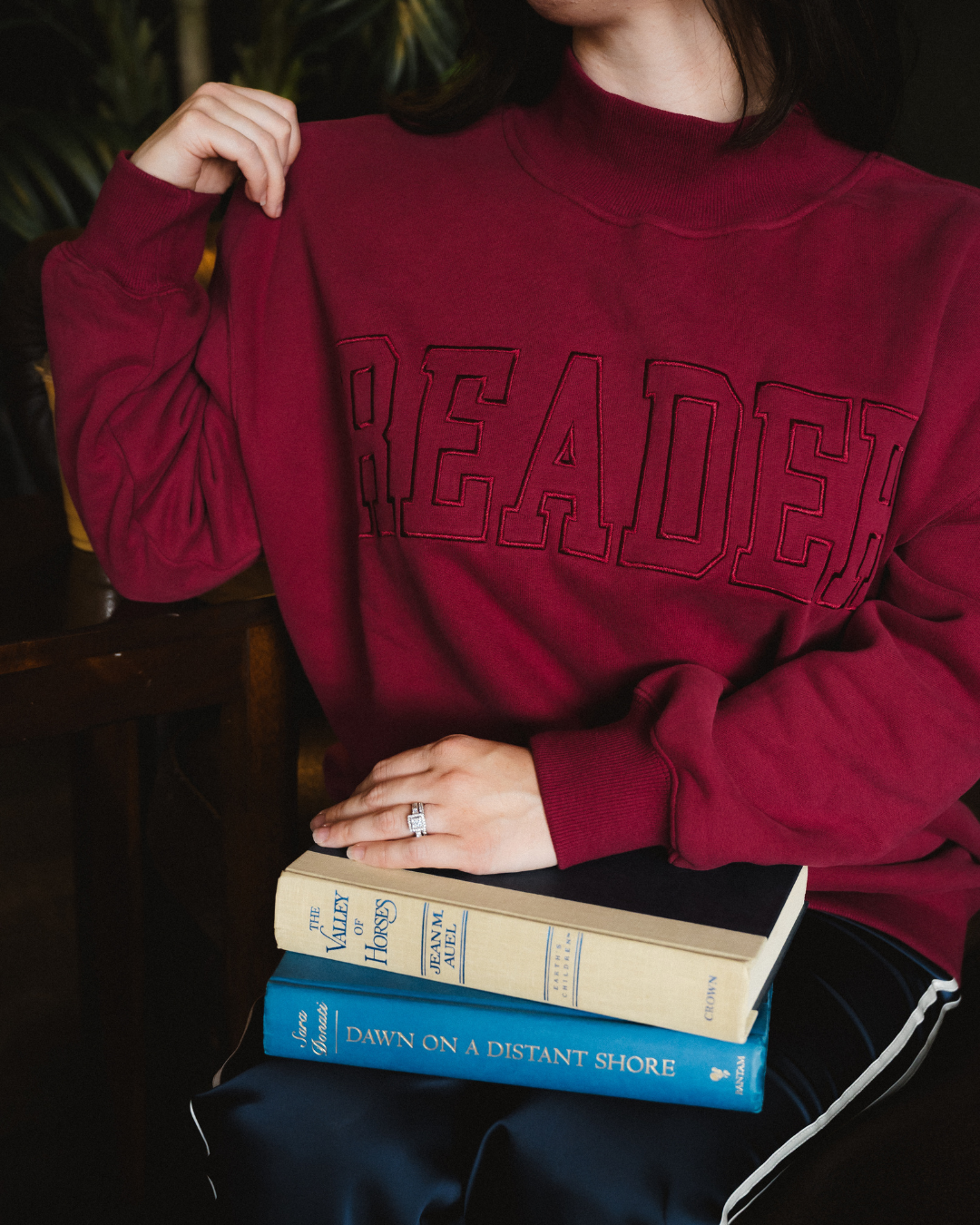 The Reader Mock Neck sweatshirt from Next Chapter is a beautiful berry, dark magenta color with an embroidered 'reader' design across the front. It's the perfect cozy bookish sweater for readers and book lovers.
