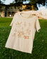 Beige t-shirt with dark brown graphic designs held by a person outdoors on grass. The design is inspired by the creatures in the Harry Potter universe.