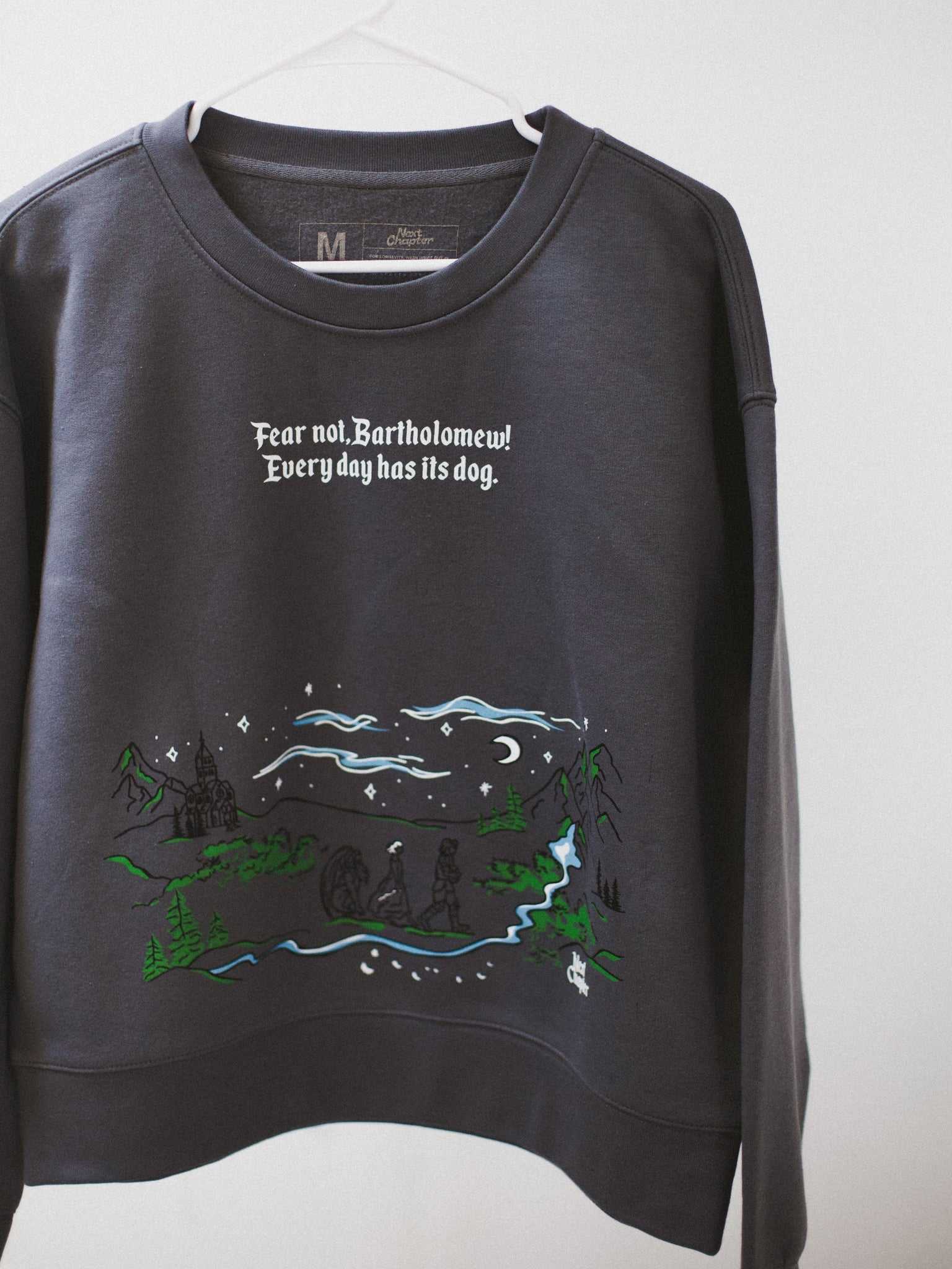 The Unlikely Trio Crewneck sweatshirt is dark gray with 'Fear not, Bartholomew! Every day has its dog' text and a colorful graphic design. Officially licensed Rachel Gillig merch. The Knight and the Moth merch. 